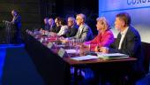 7 députés sont assis sur une scène et regardent vers le public. 