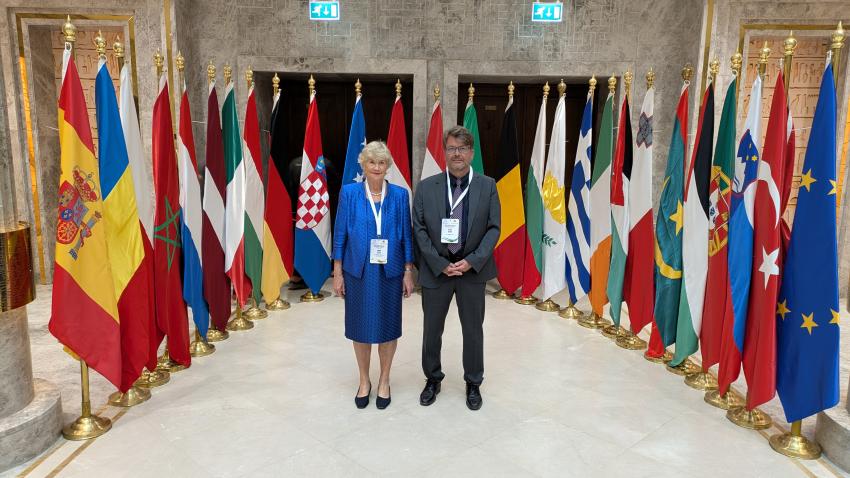 Mme Simon Beissel et M. Dan Hardy avant les drapeaux des Etats participants
