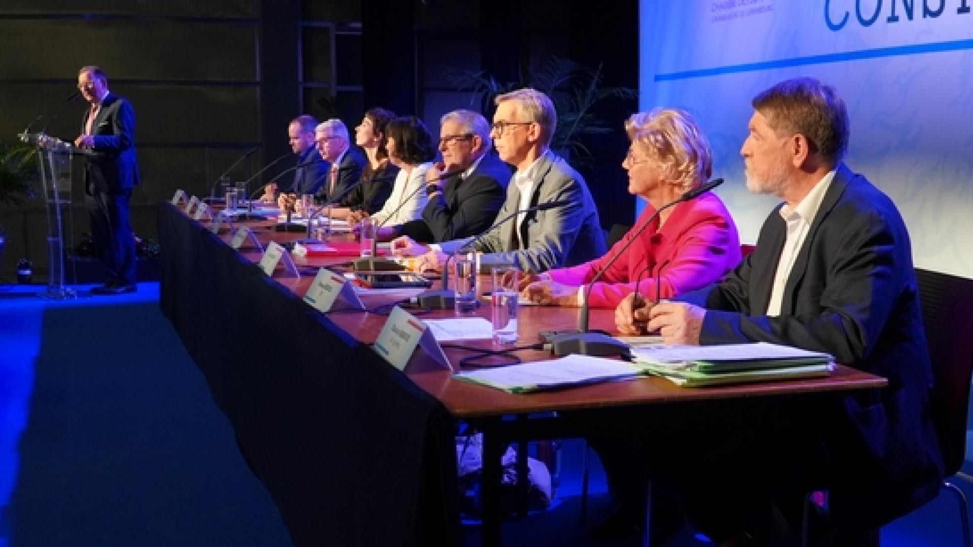 7 députés sont assis sur une scène et regardent vers le public. 